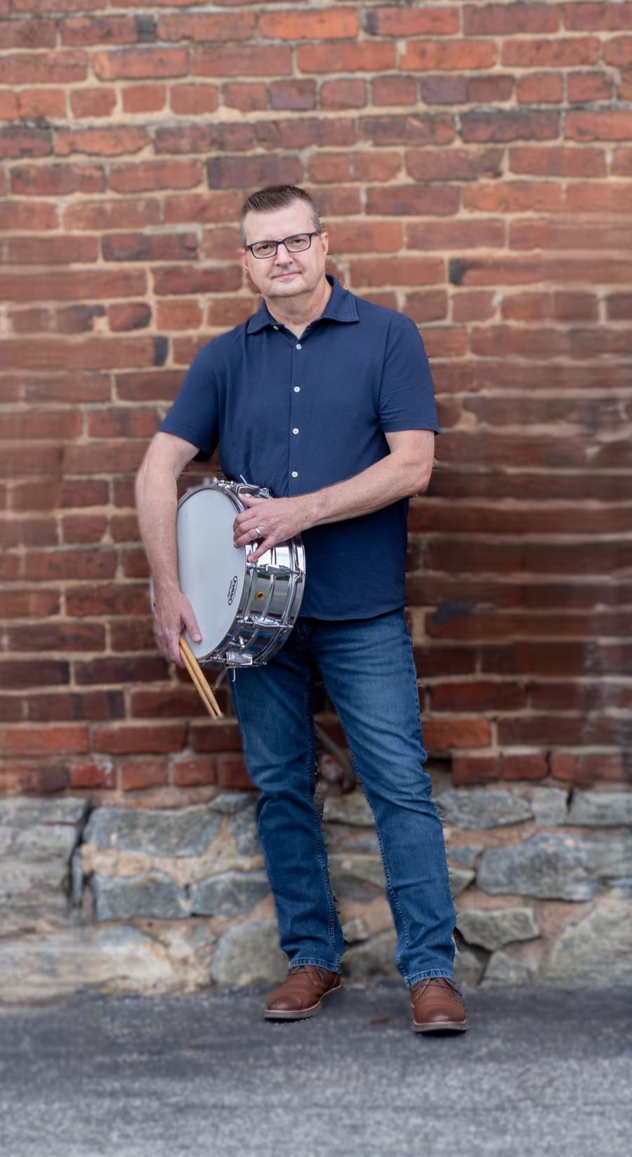 The founder holding a snare drum and sticks in front of a brick wall